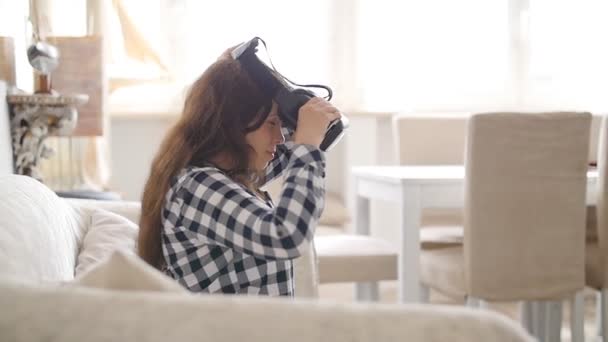 Jeune femme avec casque de réalité virtuelle ou lunettes 3D assis sur le canapé à la maison — Video