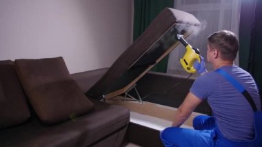 Mobilya ve apartman temizliği konsepti. Kanepeyi buharlı temizleme makinesiyle temizleyen adam.