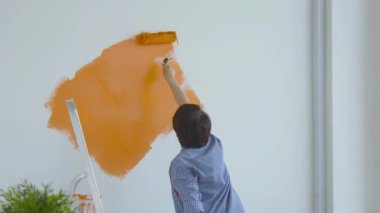 Düz yenileme konsepti. Orta yaşlı mutlu bir kadın boya silindirli, turuncu boyalı beyaz duvar boyuyor.