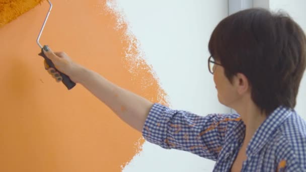 Концепция реконструкции квартир. Счастливая женщина средних лет рисует белую стену с краской роликом, оранжевой краской — стоковое видео