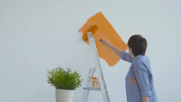 개조 개념입니다. 페인트 롤러, 오렌지 페인트로 흰색 벽을 그리는 행복 한 중년 여성 — 비디오