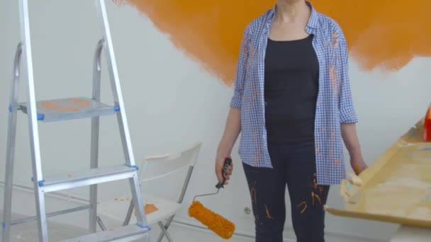 Concetto di ristrutturazione piatto. Felice donna di mezza età pittura parete bianca con rullo di vernice, vernice arancione — Video Stock