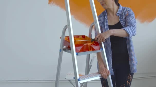 Platte renovatie concept. Gelukkig Middelbare vrouw schilderen witte muur met verfroller, oranje verf — Stockvideo