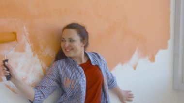 Singleton yaşam tarzı ve yenileme konsepti. Genç, gülümseyen bir kadın boya pateniyle dairedeki duvarı boyuyor ve dans ediyor.