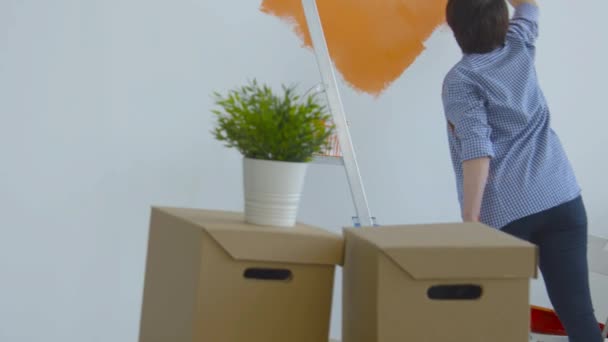 Singleton lifestyle and renovation concept. Flat renovation concept. Happy Middle-aged woman painting white wall with paint roller, orange paint — Stock Video