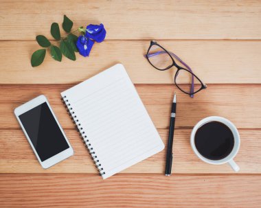 Üzerinde defter, akıllı telefon, gözlük, kahve, çiçek ve kalem olan ofis masasının üst görüntüsü..