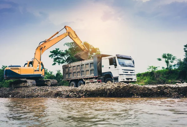 Tracked excavator loading soil and stone in a truck in the river.