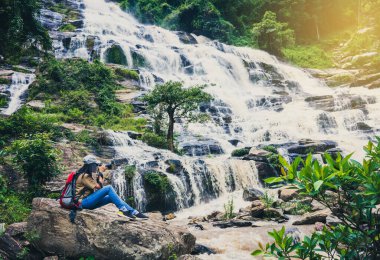 Ormanın derinliklerinde şelalenin yanında oturan Asyalı fotoğrafçı kadın fotoğrafçı kamera kullanarak fotoğraf çekiyor..