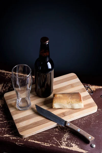 Glass and Bottle of dark beer with smoked cheese on cutting boar — Stock Photo, Image