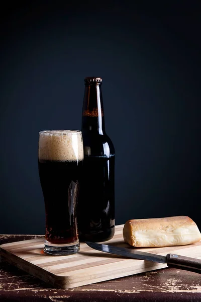 Glass and Bottle of dark beer with smoked cheese on cutting boar