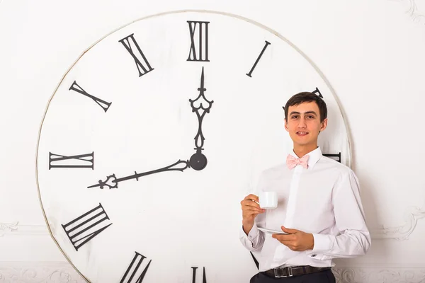 Человек с чашкой чая стоит рядом с большими часами на белой стене — стоковое фото