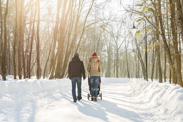 Joyeux jeune famille marchant dans le parc en hiver. Les parents portent le bébé dans une poussette à travers la neige . — Photo