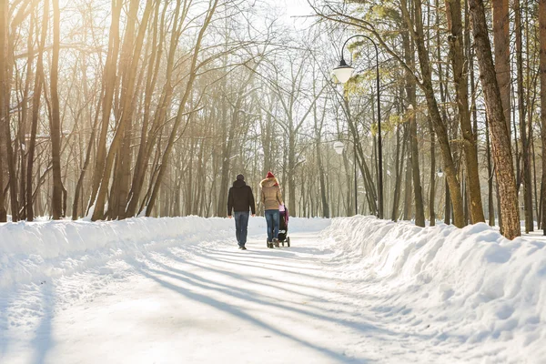Feliz jovem família caminhando no parque no inverno. Os pais carregam o bebê em um carrinho de bebê através da neve . — Fotografia de Stock