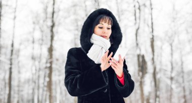 Akıllı telefonlu, kış manzaralı ve arka planda kar taneleriyle gülümseyen genç bir kadın.