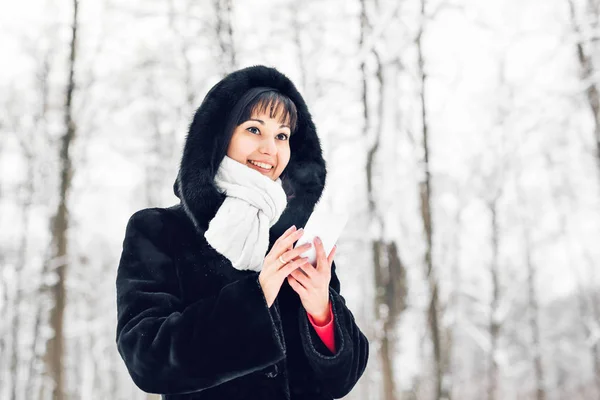 Fiatal nő mosolyog okos telefon és téli táj és hópelyhek a háttérben — Stock Fotó
