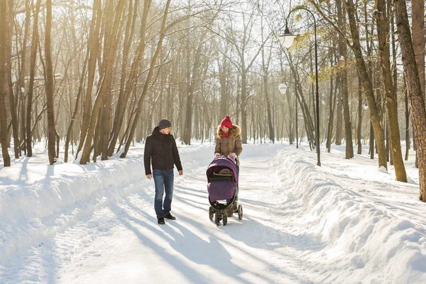 Feliz jovem família caminhando no parque no inverno. Os pais carregam o bebê em um carrinho de bebê através da neve . — Fotografia de Stock