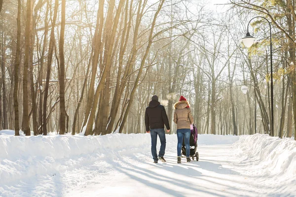 Joyeux jeune famille marchant dans le parc en hiver. Les parents portent le bébé dans une poussette à travers la neige . — Photo