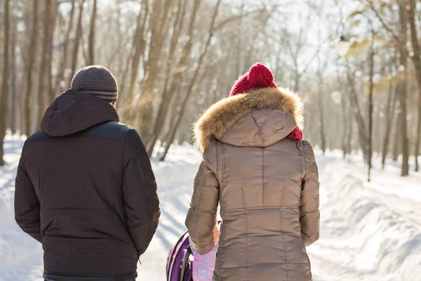 Joyeux jeune famille marchant dans le parc en hiver. Les parents portent le bébé dans une poussette à travers la neige . — Photo