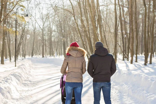 Glad ung familj promenader i parken på vintern. Föräldrarna bär barnet i en barnvagn genom snön. — Stockfoto