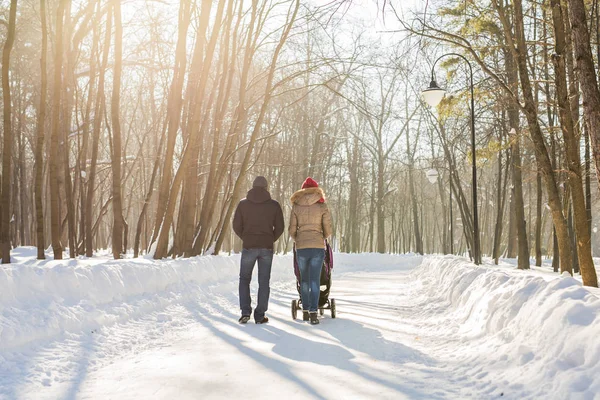 Joyeux jeune famille marchant dans le parc en hiver. Les parents portent le bébé dans une poussette à travers la neige . — Photo