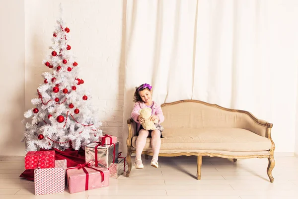Smiling kid girl holding white teddy bear with christmas tree indoors. Childhood. — Stock Photo, Image