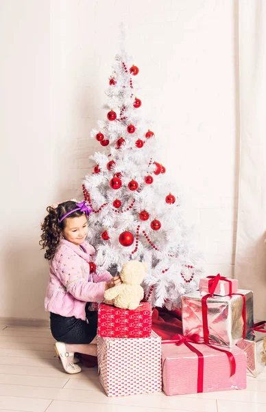 Niña sonriente sosteniendo oso de peluche blanco con árbol de Navidad en el interior. Infancia . — Foto de Stock