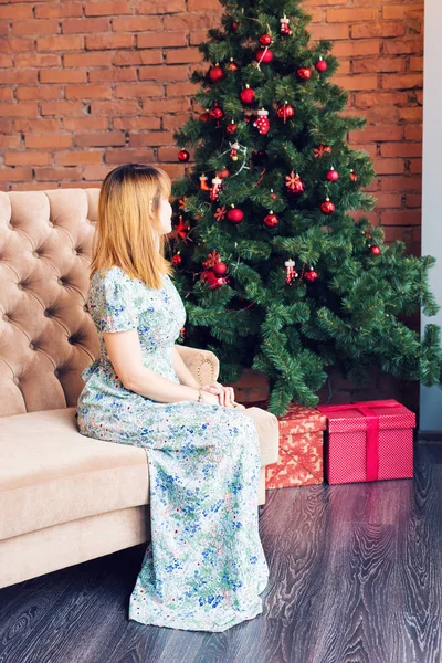 Eine junge Frau sitzt auf einem Sofa neben einem Weihnachtsbaum mit Geschenkschachteln. Neujahrs- und Weihnachtskonzept. — Stockfoto