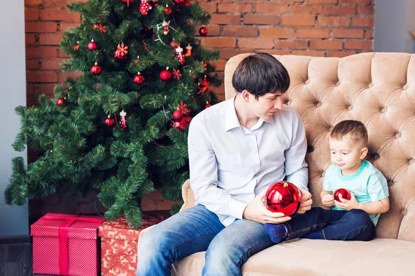 Jovem pai brincando com seu filho bebê no sofá perto da árvore de Natal — Fotografia de Stock