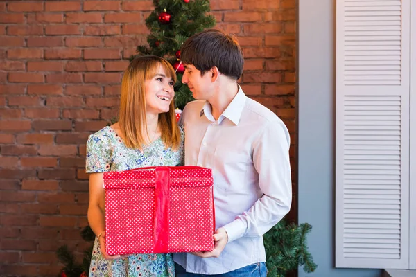 Pareja compartiendo regalo de Navidad. Joven familia abrazando y sosteniendo caja de regalo roja en casa con decoraciones — Foto de Stock