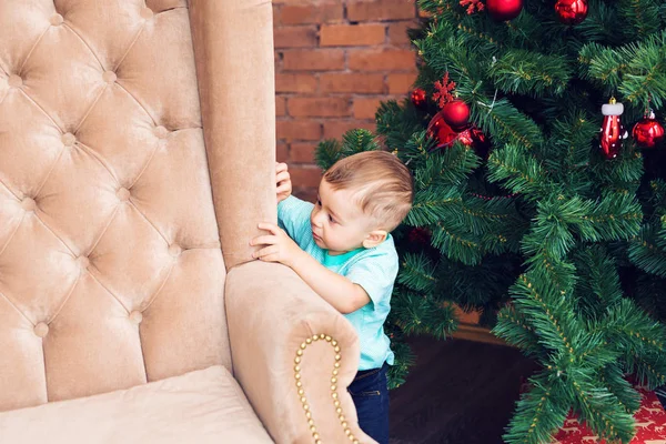 Divertido niño de pie cerca del sofá y el árbol de Navidad con colorida bola de Navidad — Foto de Stock
