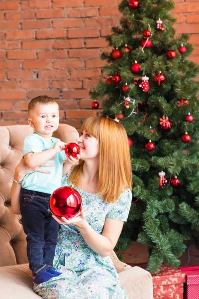 Retrato de madre feliz y bebé riendo sosteniendo chuchería contra el interior festivo doméstico con árbol de Navidad . — Foto de Stock