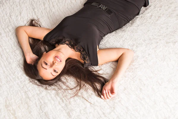 Mooie en aantrekkelijke jonge vrouwelijke brunette sensualiteit vrouw poseren in zwarte jurk op bed in appartement — Stockfoto