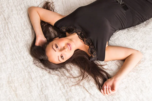 Mulher elegante em vestido preto moda lindo com penteado maquiagem brilhante no apartamento do hotel. Sexy menina magro relaxante na cama em casa interior de luxo. Modelo de moda . — Fotografia de Stock