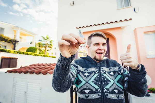 Portrait of a young happy man holding out new house key and gesturing thumbs up