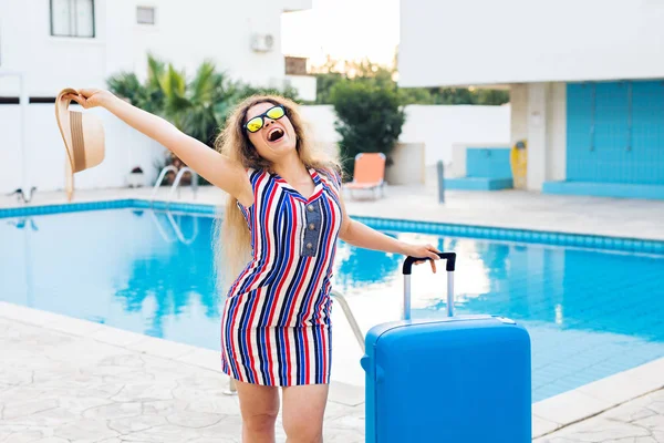Gelukkig jonge vrouw met blauwe bagage aankomen aan het resort. Ze loopt naast het zwembad. Begin van de zomer vakantie concept. — Stockfoto