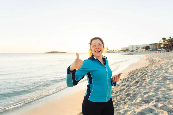 Concept van sport, fitness, gezonde levensstijl en running - gemotiveerd sportieve vrouw doen duimschroef opwaarts succes gebaar na buiten training op het strand — Stockfoto