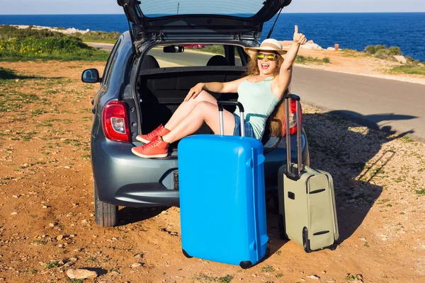 Viajes, vacaciones de verano y el concepto de vacaciones - Mujer joven con maletas en viaje en coche. Ella está sentada en el coche hacia atrás y gesto pulgares hacia arriba . —  Fotos de Stock