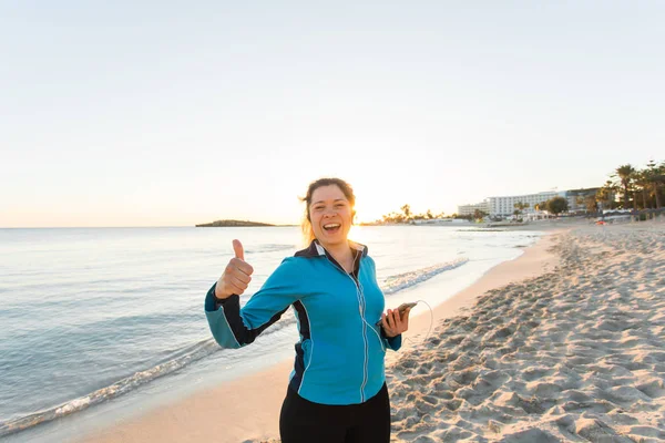Buitensport, fitness gadget en mensen concept - glimlachend vrouwelijke fitness Toon duimen omhoog ingedrukt te smartphone met koptelefoon. — Stockfoto