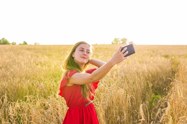 Tecnología, vacaciones de verano, vacaciones y el concepto de la gente - sonriente mujer joven tomando selfie por teléfono inteligente en el campo de cereales . — Foto de Stock