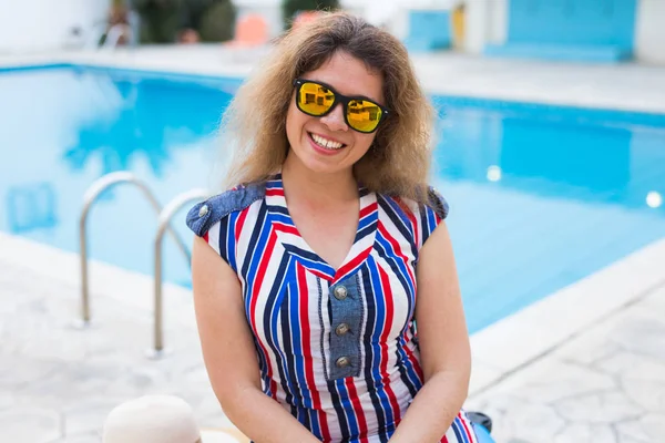 Portret van gelukkig meisje op zomervakantie op de achtergrond van zwembad, gekleed gestreepte jurk en zonnebril. — Stockfoto