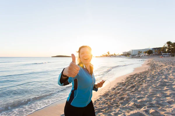 Mujer deportiva motivada haciendo pulgares gesto de éxito después del entrenamiento urbano en la orilla del mar . — Foto de Stock