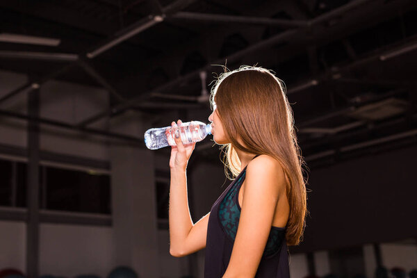 Fitness woman drinking water from bottle. Muscular young female at gym taking a break from workout.