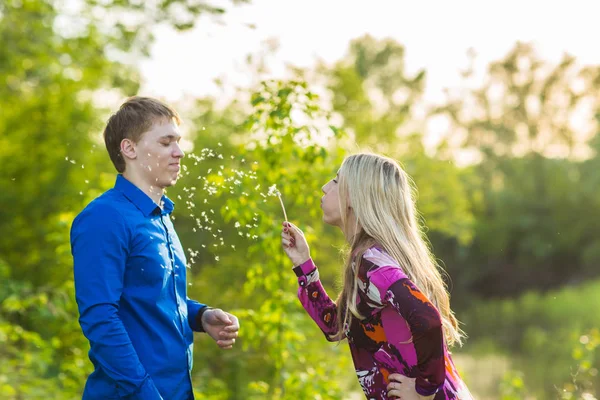 Par i kärlek blåser blowballs blommor i ansiktet på varandra. Ler och skrattar folk har god tid utanför varm sommardag. Kvinna och man njuter av naturen. — Stockfoto