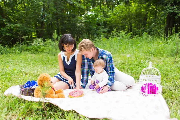 Jovem mãe e pai felizes com sua filha bebê relaxando em um cobertor em um parque comemorando com bolo de aniversário — Fotografia de Stock