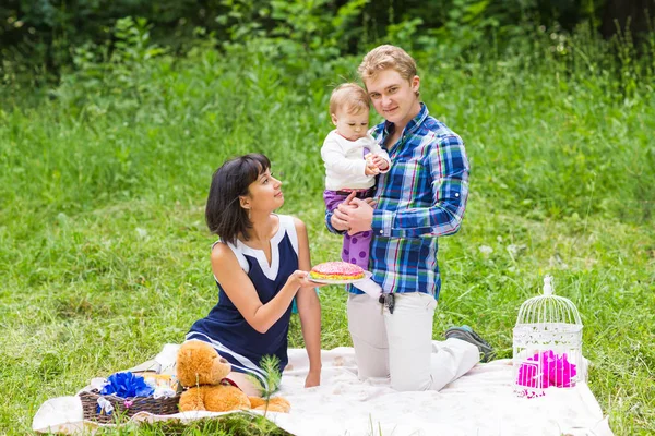 Szczęśliwa młoda matka i ojciec z córką baby relaksujący na kocu w parku obchodzi z tort urodzinowy — Zdjęcie stockowe