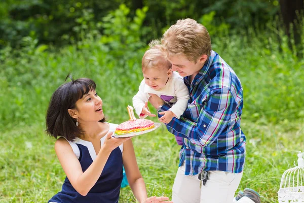 Jovem mãe e pai felizes com sua filha bebê relaxando em um cobertor em um parque comemorando com bolo de aniversário — Fotografia de Stock