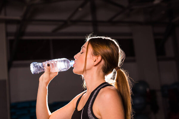 Fitness woman drinking water from bottle. Muscular young female at gym taking a break from workout.