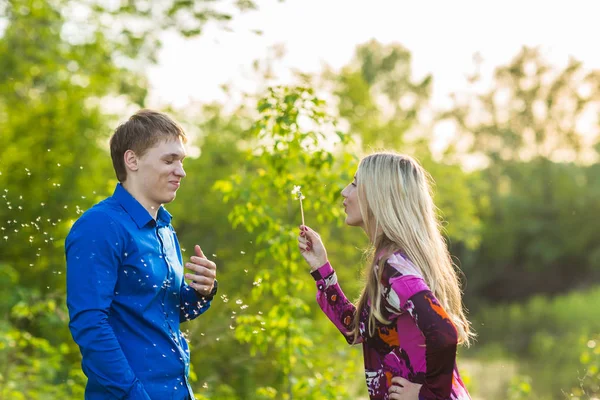 Par i kärlek blåser blowballs blommor i ansiktet på varandra. Ler och skrattar folk har god tid utanför varm sommardag. Kvinna och man njuter av naturen. — Stockfoto