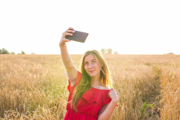 Technologie, letní prázdniny, dovolené a lidé koncept - usmívající se dívka v červených šatech pořizování selfie pomocí smartphone na obilné pole — Stock fotografie
