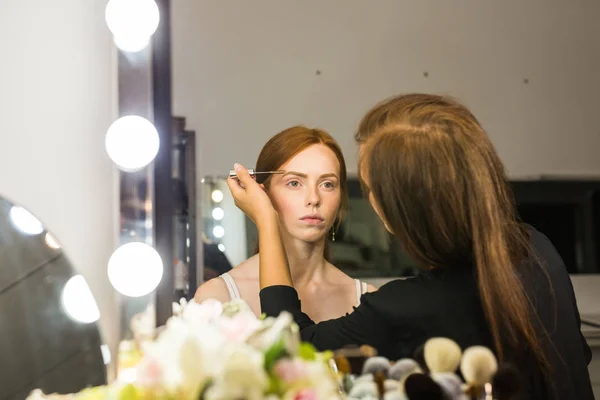 Prozess der Herstellung von Make-up. Make-up-Artist arbeitet mit Pinsel auf Modell Gesicht. Porträt einer jungen Frau mit rotem Haar im Interieur eines Schönheitssalons. Ton auf die Haut auftragen. — Stockfoto